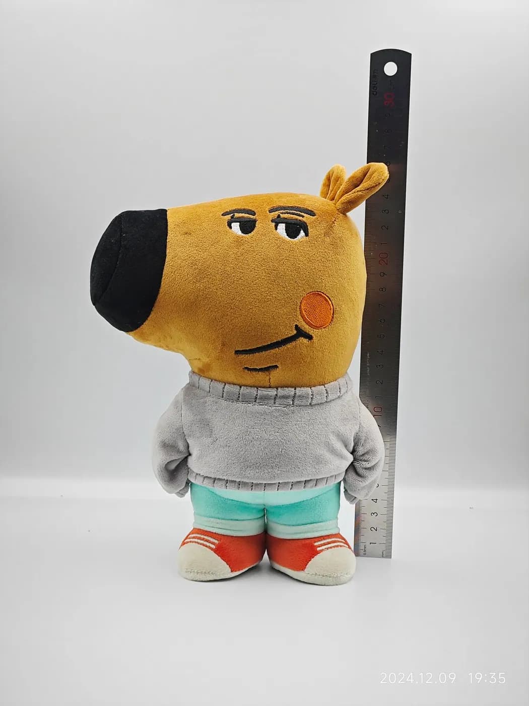 Orange dog character plush in a knitted jumper, measured against a steel rule